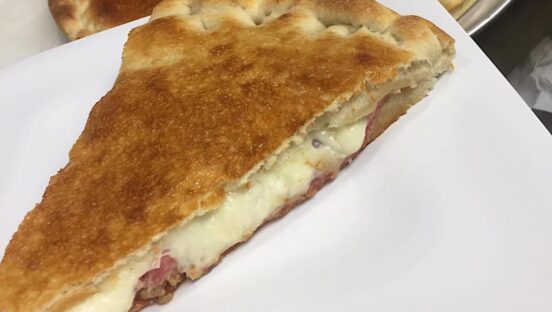 This photo shows a pizza stuffed with six meats and three layers of cheese on a paper plate.
