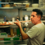 This photo shows Jose Rodriguez, wearing a light green T-shirt, peeling a pizza into his oven at Brooklyn Pizza.