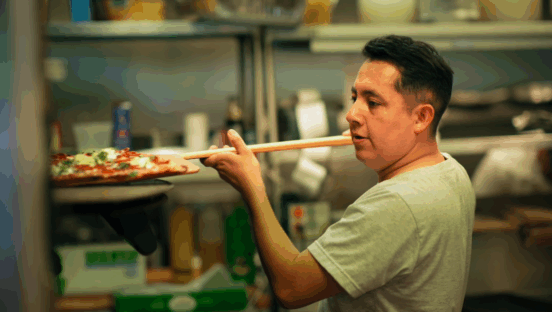 This photo shows Jose Rodriguez, wearing a light green T-shirt, peeling a pizza into his oven at Brooklyn Pizza.