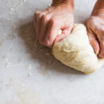 This photo shows a man's hands kneading pizza dough on a white table.