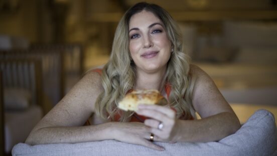 This photo shows Erica D'arcangelo smiling with a slice of pizza while leaning against the top of a gray sofa.
