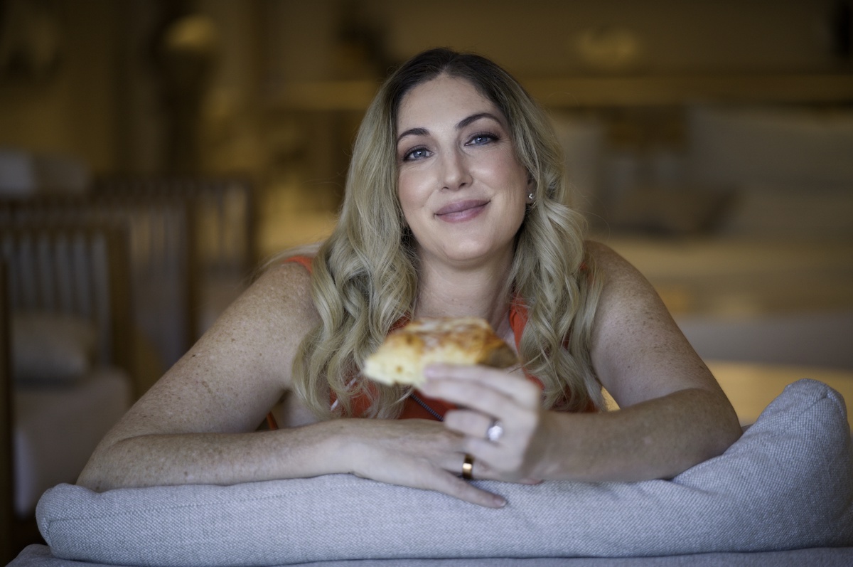 This photo shows Erica D'arcangelo smiling with a slice of pizza while leaning against the top of a gray sofa.