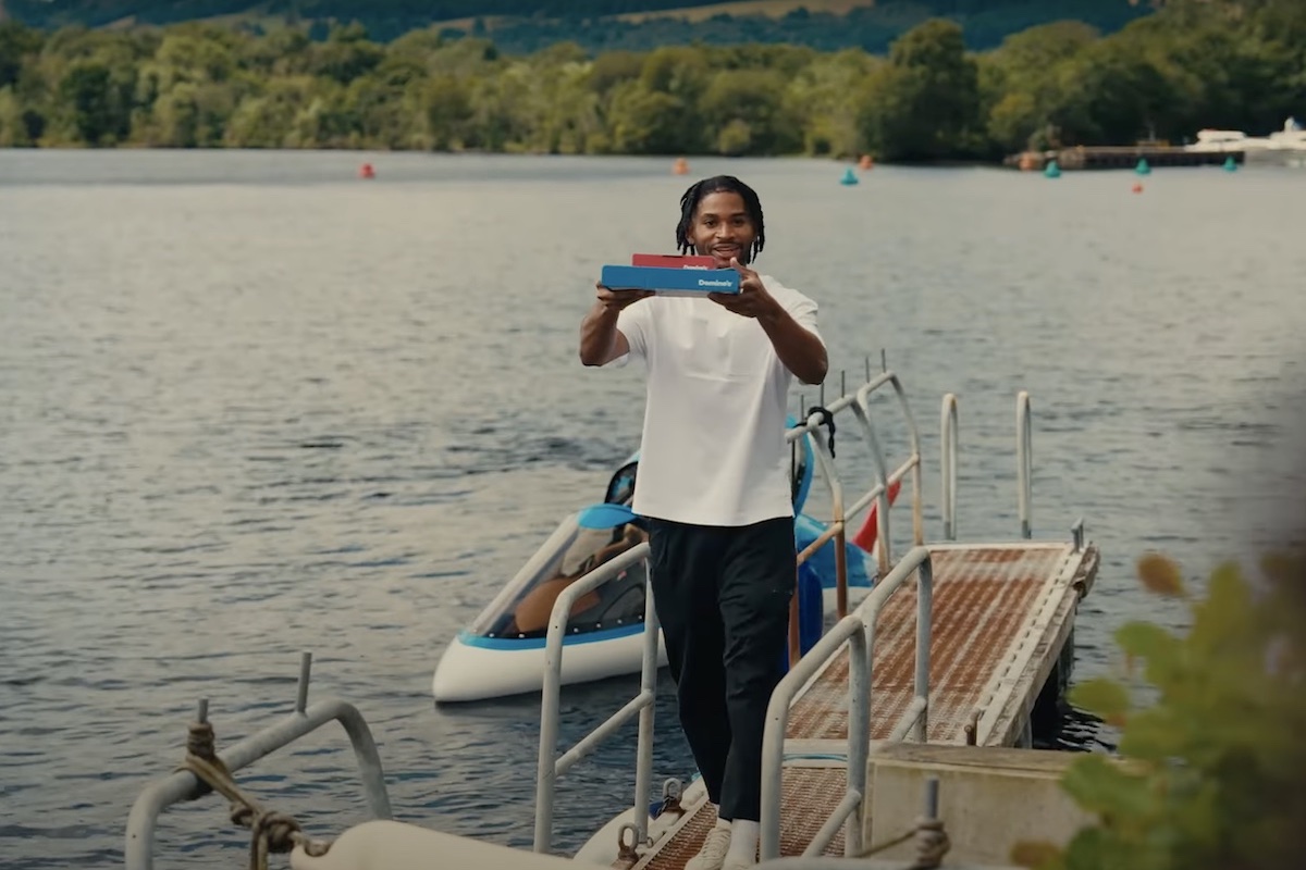 This photo shows a man holding up a pizza box on the dock at Loch Ness while a submarine waits on the water behind him.