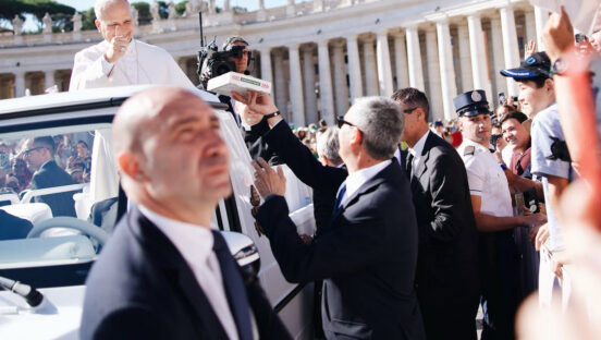 This photo, shot from a distance, shows a security agent passing a pizza box to a white-clad Pope Leo XIV in the popemobile on St. Peter's Square.