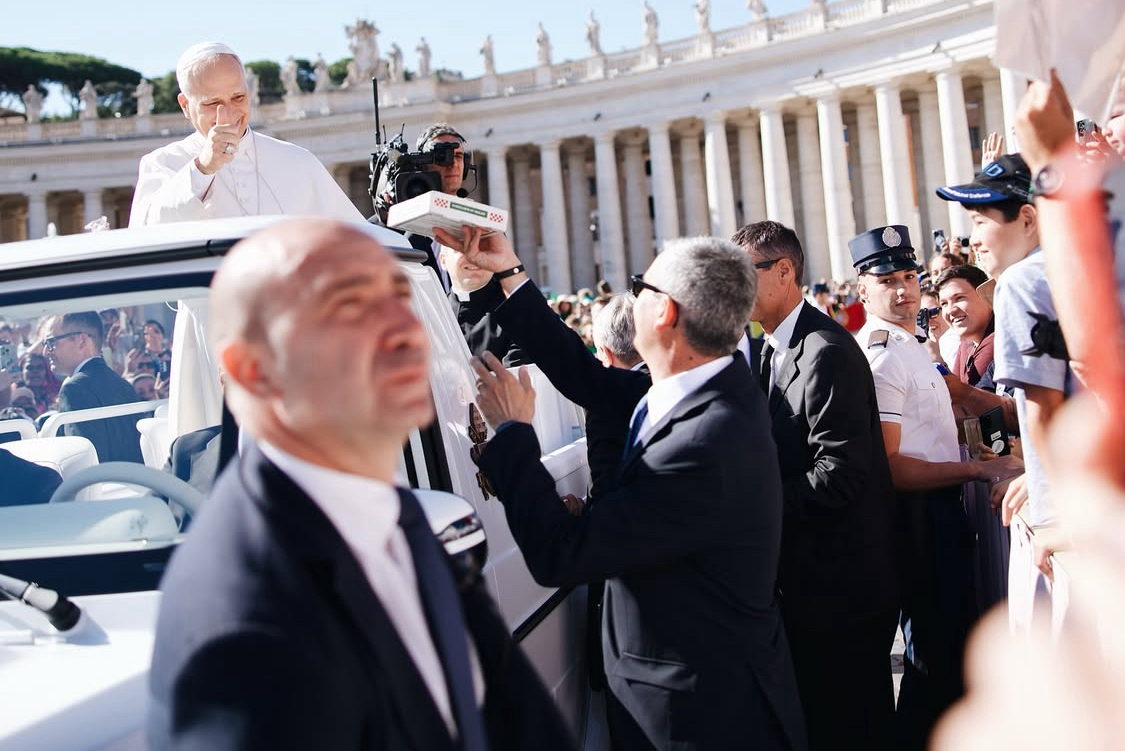 This photo, shot from a distance, shows a security agent passing a pizza box to a white-clad Pope Leo XIV in the popemobile on St. Peter's Square.