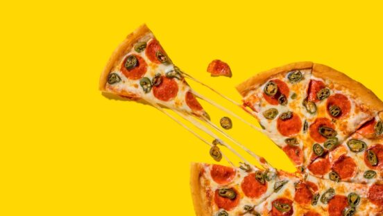 This photo shows a slice pulled out from a whole pie topped with pepperoni and jalapeños, set against a yellow background.