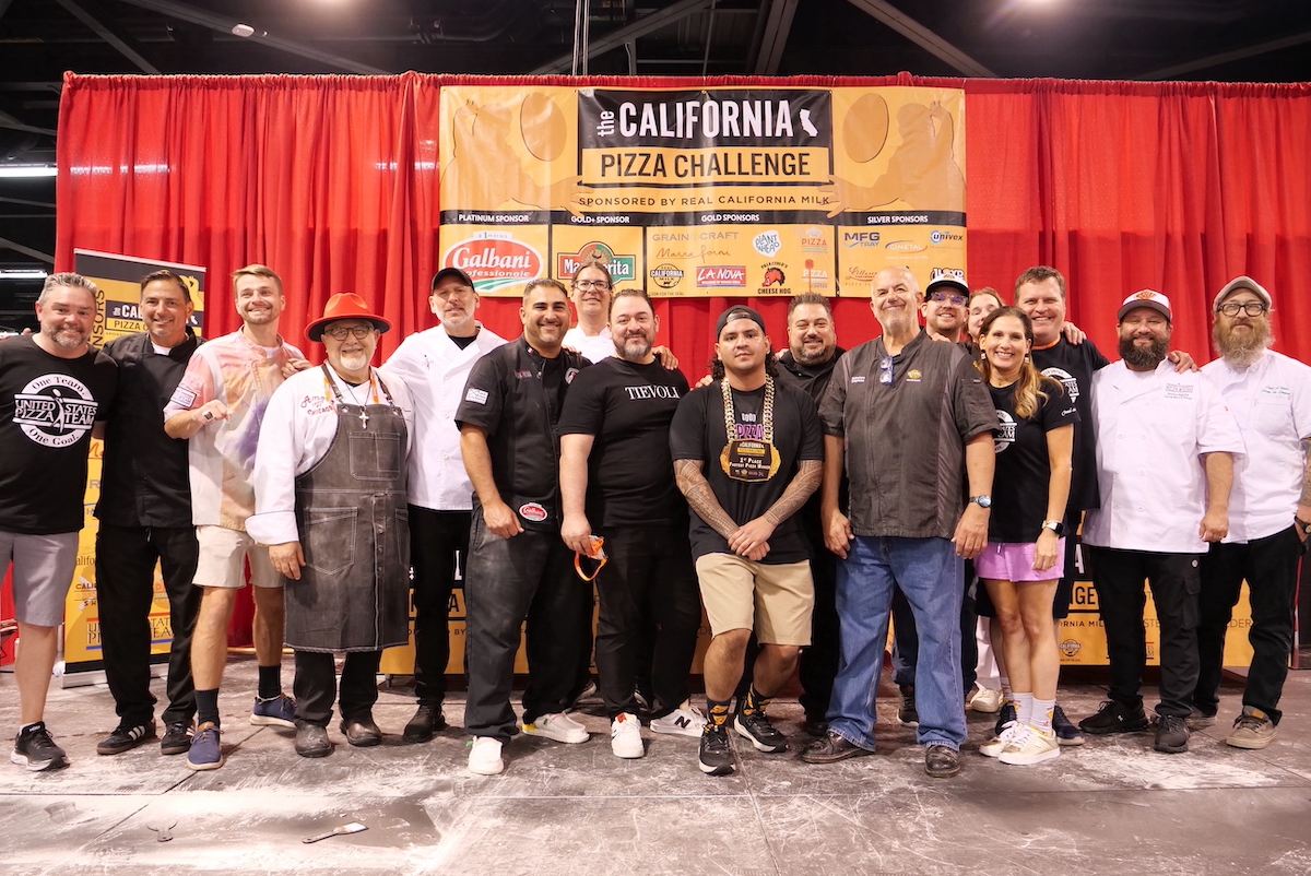 This photo shows numerous members of the U.S. Pizza Team standing together at a competition event in front of a red backdrop.