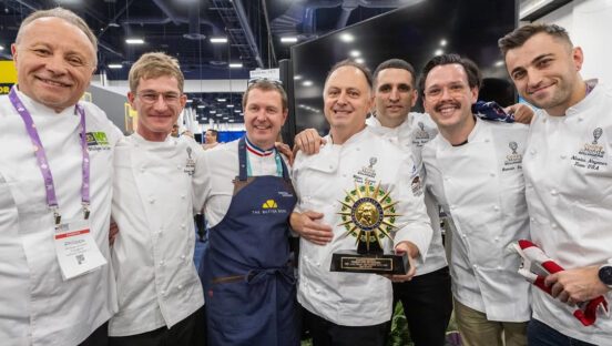 This photo shows members of Team USA, all wearing white chef coats, at the IBIE competition. The man in the middle is holding the 2nd place trophy.