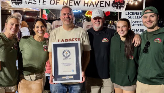 This photo shows Colin Kaplan holding a framed Guinness World Records certificate. He is flanked by Big Green Truck Pizza owner Liane Page on his right as well as her family members and employees.