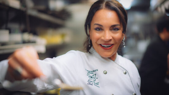 This photo shows Lisa Dahl presenting what appears to be a spoon with sauce in it (blurred at foreground) toward the camera.