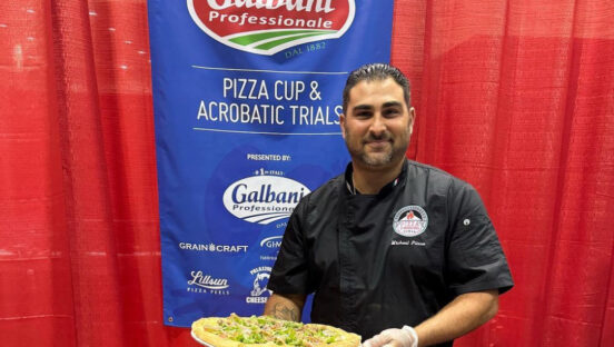 This photo shows Mike Pitera holding a pizza at a culinary competition. He is standing in front of a banner for Galbani Professionale.