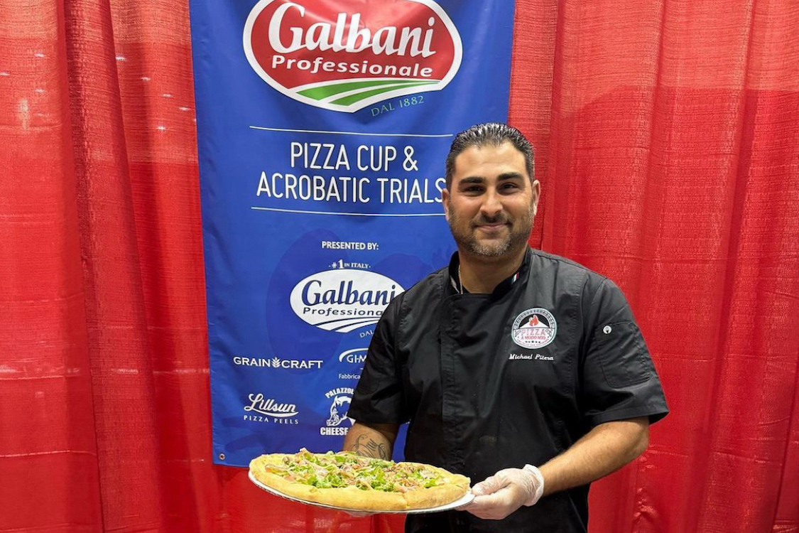 This photo shows Mike Pitera holding a pizza at a culinary competition. He is standing in front of a banner for Galbani Professionale.