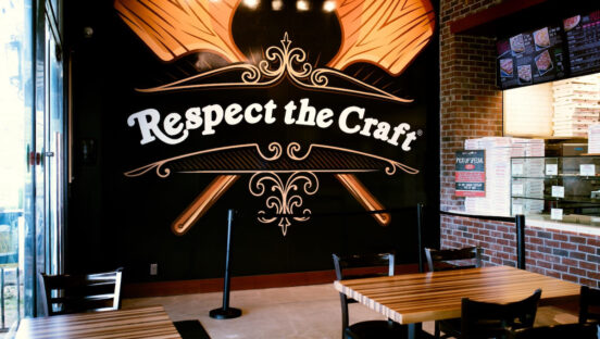 This photo shows the interior of a Slice House restaurant, centered on a large artwork depicting two pizza peels crossed like an X and the words Respect the Craft.