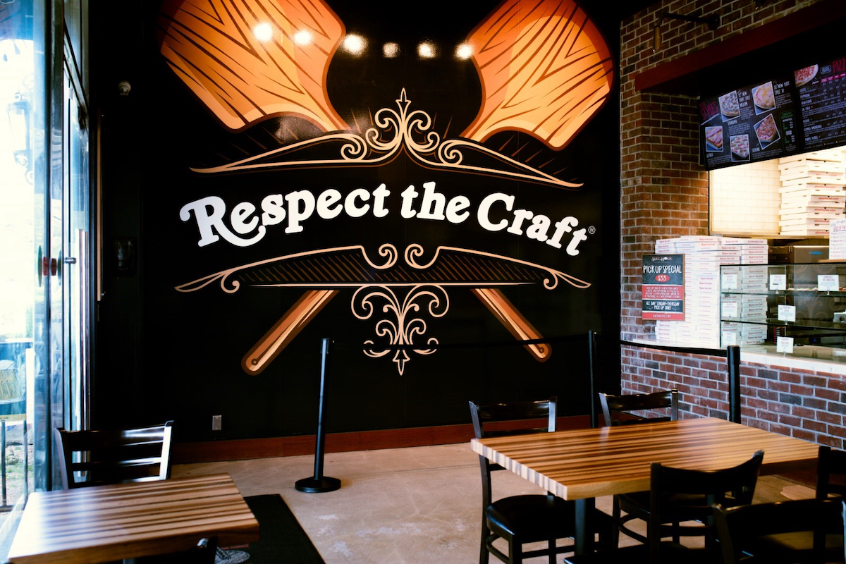 This photo shows the interior of a Slice House restaurant, centered on a large artwork depicting two pizza peels crossed like an X and the words Respect the Craft.