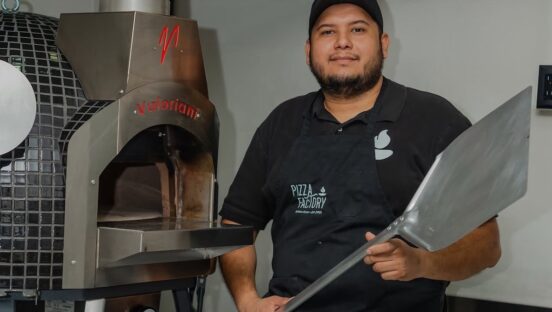 This photo shows Antonio Portillo in a black Pizza Factory-branded shirt, holding a metal pizza peel and standing in front of his pizza oven.