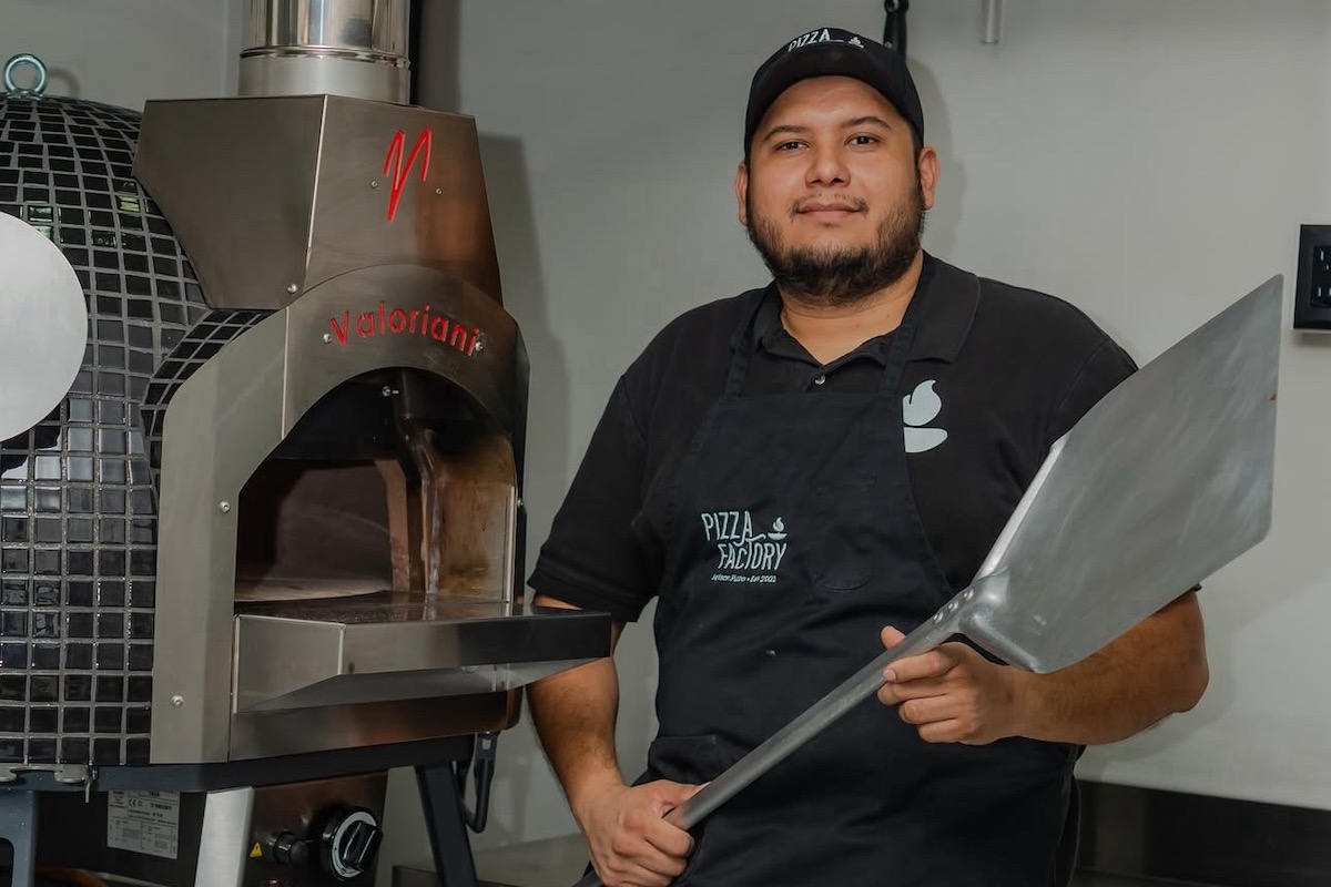 This photo shows Antonio Portillo in a black Pizza Factory-branded shirt, holding a metal pizza peel and standing in front of his pizza oven.