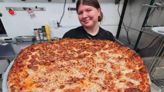 This photo shows an employee of Circles & Squares holding up a huge oversized round pizza with half cheese and half pepperoni.