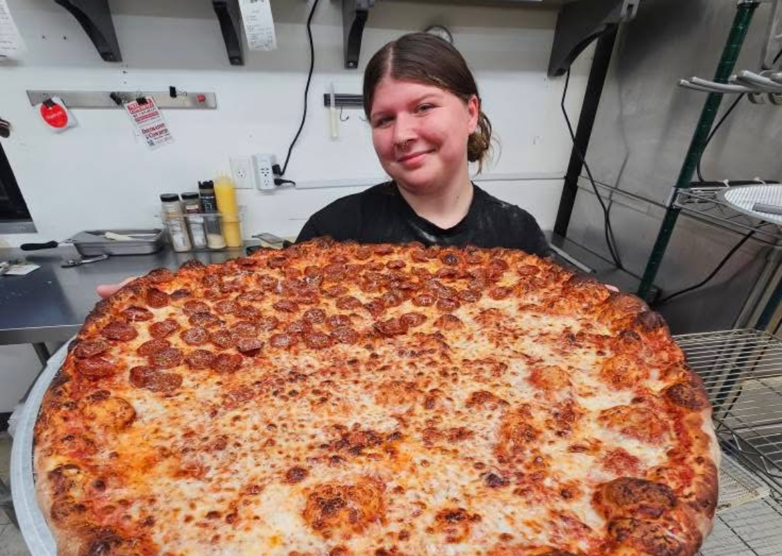 This photo shows an employee of Circles & Squares holding up a huge oversized round pizza with half cheese and half pepperoni.