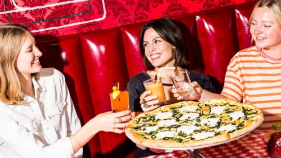This photo shows three female customers clinking their drinks together before digging into a large pizza on the table in front of them at Home Slice Pizza.