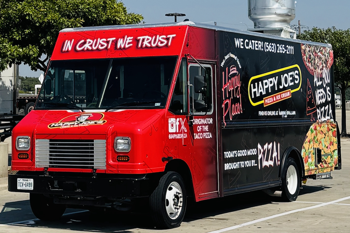 This photo shows the Happy Joe's Pizza food truck, bright red on the front with "In Crust We Trust" in white lettering above the windshield and a black side panel that's branded with various mottos, such as Today's Good Mood Brought to You by...Pizza."
