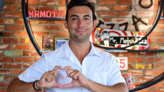 This photo shows Gibran Fernandez, wearing a white shirt, making a heart sign with his fingers over the Mr. Moto logo on the front of the shirt.