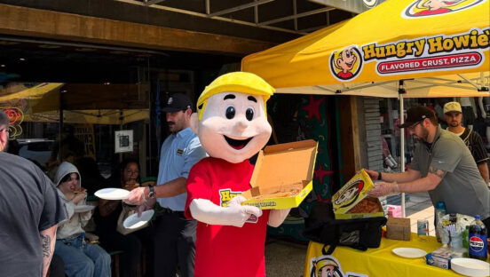 This photo shows the Hungry Howie's blonde-haired mascot in a red t-shirt standing next to a Hungry Howie's pizza stand in front of a tattoo parlor.