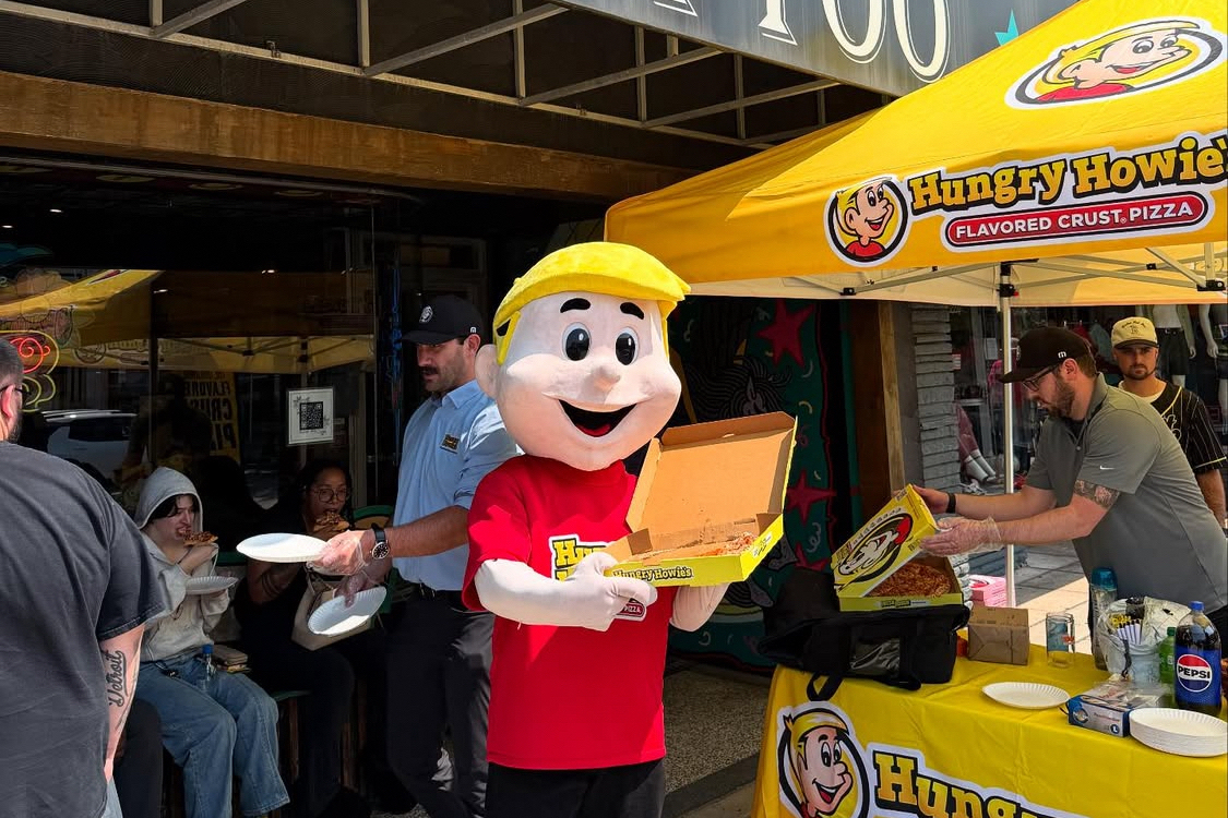This photo shows the Hungry Howie's blonde-haired mascot in a red t-shirt standing next to a Hungry Howie's pizza stand in front of a tattoo parlor.
