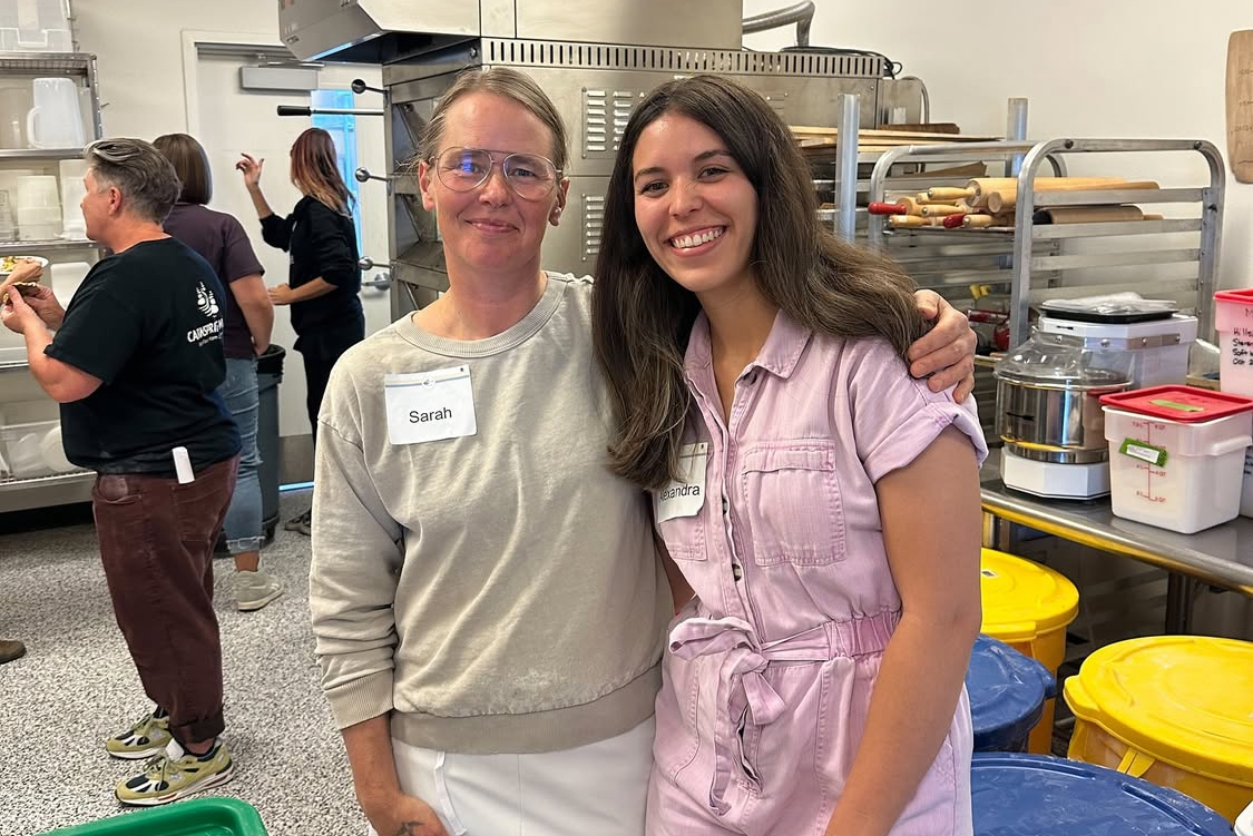 This photo shows Alexandra Castro standing at left, wearing a pink dress, with Sarah Minnick, wearing a gray sweater, standing together and smiling for the camera.