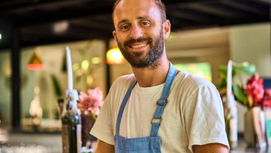 This photo shows Sebastian Galluci, wearing a white t-shirt and a blue apron, standing in his restaurant.