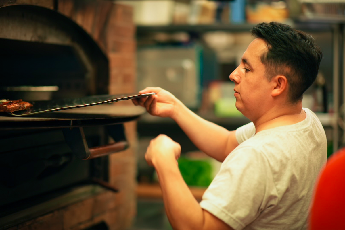 This photo shows Jose Rodriguez, wearing a white t-shirt, placing a pizza into his oven.