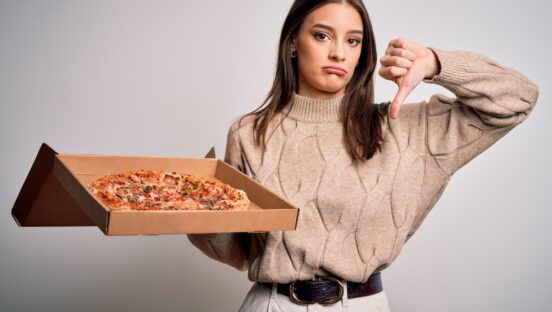 This photo shows an unhappy-looking young woman in a brown sweater. She's holding a delivered pizza in one hand and giving the thumbs-down sign with the other.