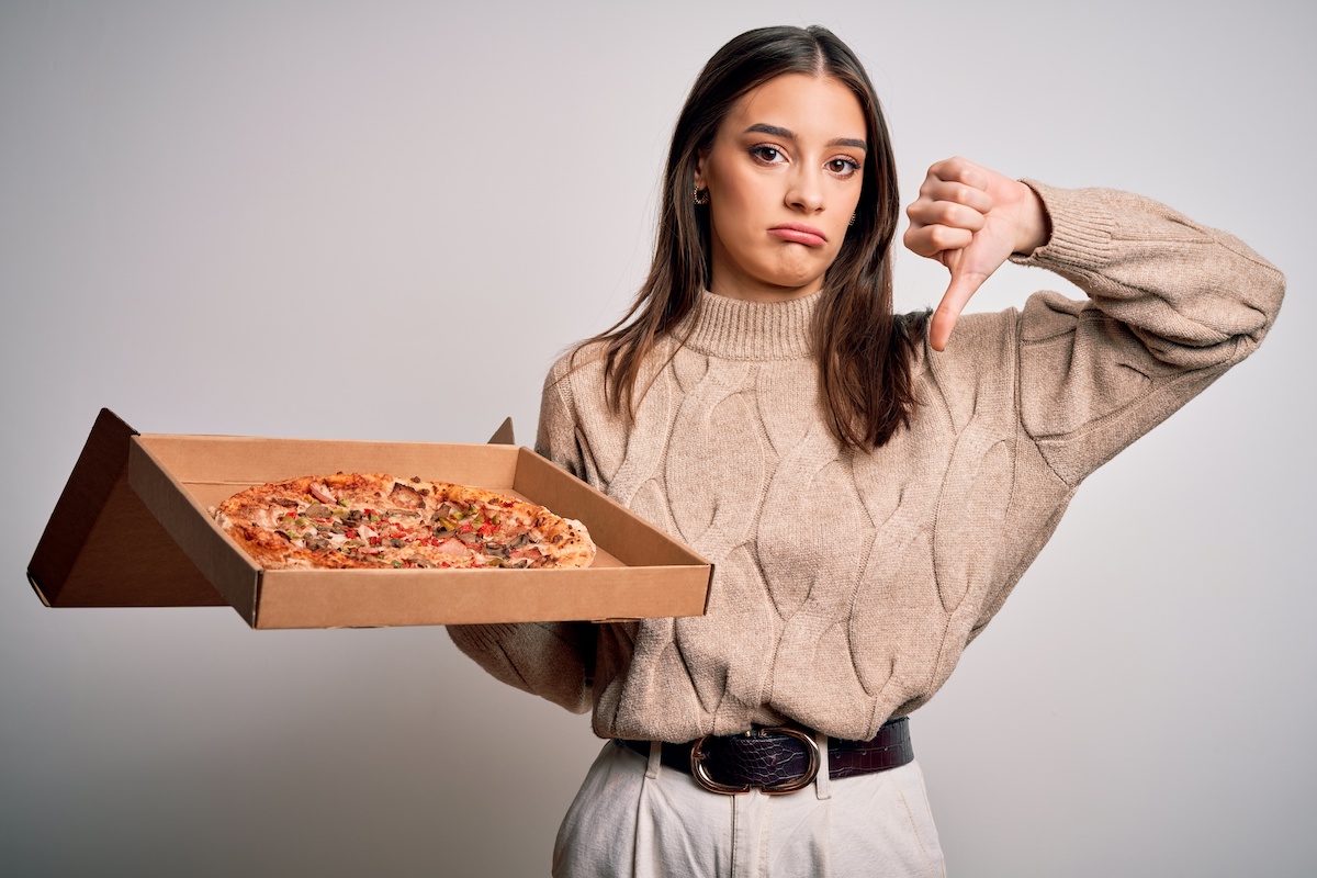 This photo shows an unhappy-looking young woman in a brown sweater. She's holding a delivered pizza in one hand and giving the thumbs-down sign with the other.