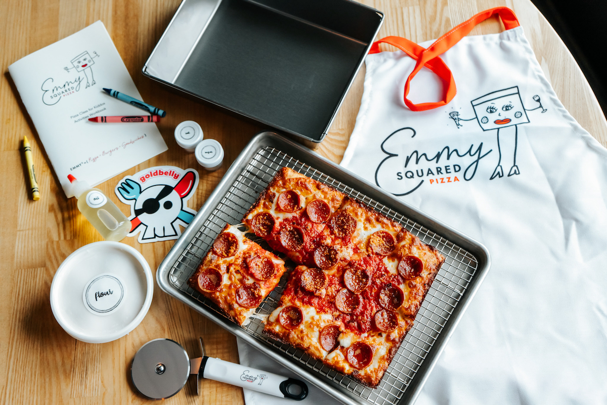 This photo shows a whole Detroit-style pizza in a pan, with an Emmy Squared apron, an Emmy Squared coloring book, another Detroit-style pan and a Goldbelly sticker.