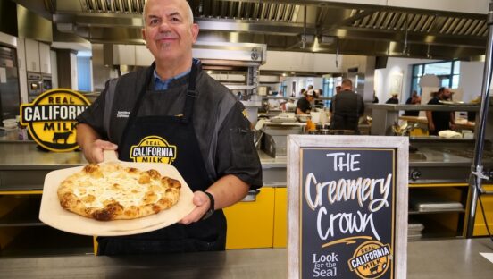 This photo shows Tore Trupiano, wearing a black chef coat, holding his Creamery Crown pizza and standing next to a sign with the pizza's name in cursive letters.