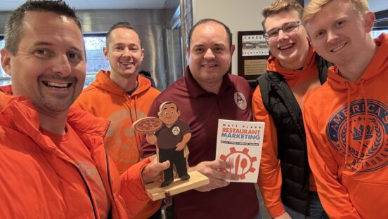 This photo shows Matt Plapp and members of his team, all wearing orange jackets, with Aldo Salazar in the center. Aldo is holding Matt's book, "Restaurant Marketing That Works."