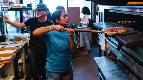 A young woman in a blue t-shirt peels a baked pizza out of the oven in Fine Folks Pizza's kitchen.