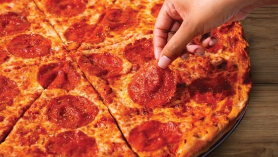 This photo shows a person's hand pulling a slice of pepperoni off a thin-crust pizza.