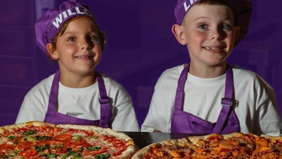 This photo shows Willow and Liam Glynn in purple chef hats and aprons smiling and posing with their pizzas against a purple background.