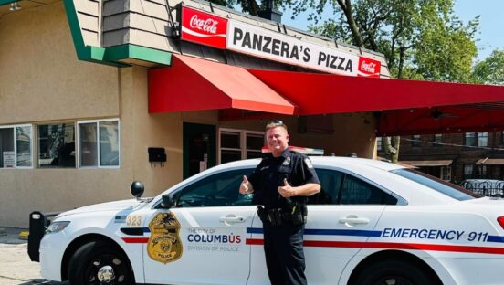 This photo shows a police officer, standing next to his squad car, giving the thumbs-up in front of Panzera's Pizza.