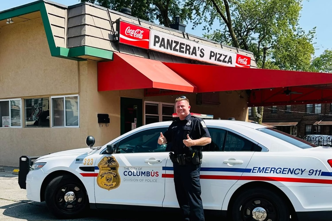 This photo shows a police officer, standing next to his squad car, giving the thumbs-up in front of Panzera's Pizza.