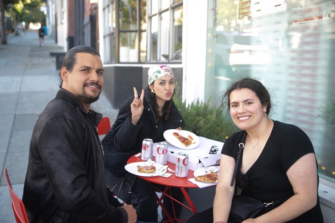 This photo shows three customers - one male and two female - eating at a table outside Goat Hill Pizza. One of the women is flashing a peace sign with her fingers.