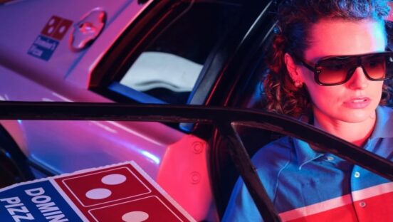 This highly stylized photo shows a woman, wearing sunglasses, getting out of a flashy white car with a pizza box. She is dressed in a Domino's uniform and looks to be a model rather than an actual delivery person.