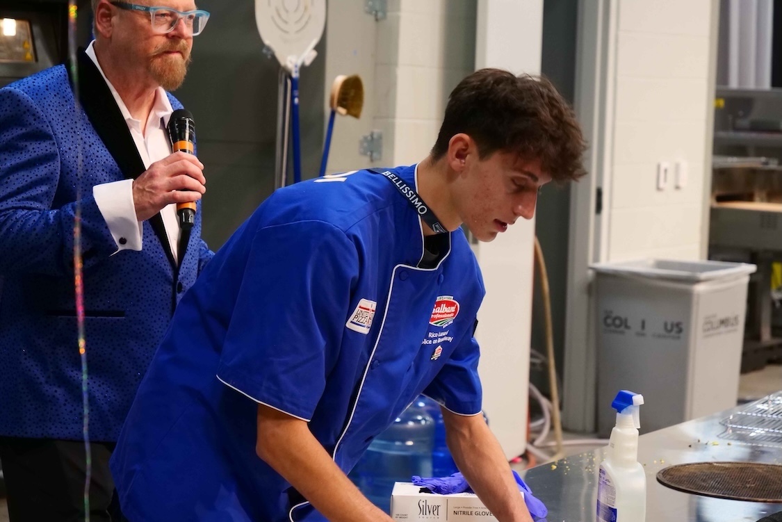 This photo shows a male competitor, wearing a blue chef coat, making a pizza at the competition described in the story.
