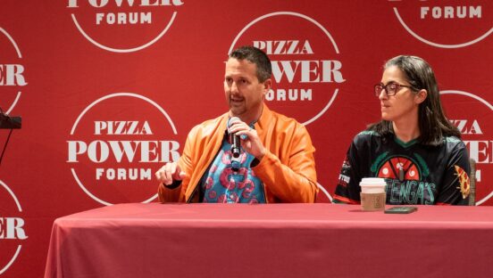 This photo shows Matt Plapp, wearing his trademark orange jacket, sitting at a table with Hengam Stanfield during a presentation at the Pizza Power Forum.