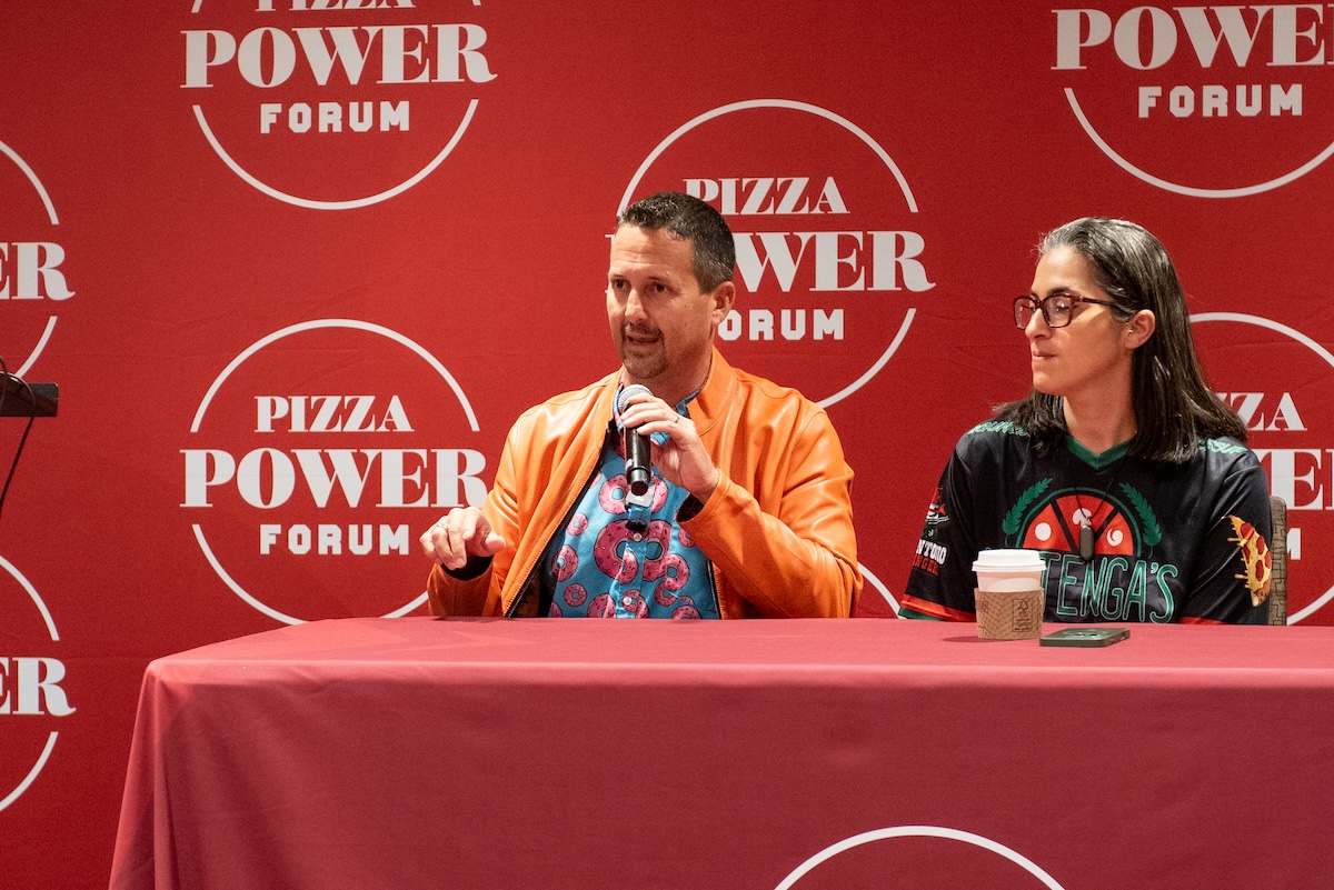 This photo shows Matt Plapp, wearing his trademark orange jacket, sitting at a table with Hengam Stanfield during a presentation at the Pizza Power Forum.
