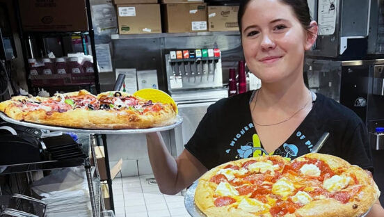 This photo shows a young, dark-haired woman in a black t-shirt holding up two different pizzas at a Mellow Mushroom location.