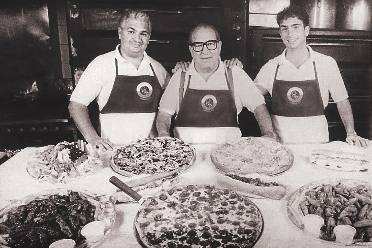 This black-and-white photo shows three of the Todaro men standing in front of a table loaded with various whole pizzas.