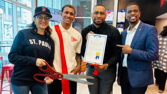 This photo shows the owners of Slice Brothers standing in the middle. On the left is a woman holding a pair of giant scissors for a ribbon-cutting ceremony. On the right is the mayor of Minneapolis.