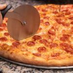This photo shows a New York-style pepperoni pizza being sliced by a metal slicer.