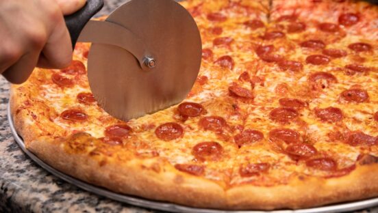 This photo shows a New York-style pepperoni pizza being sliced by a metal slicer.